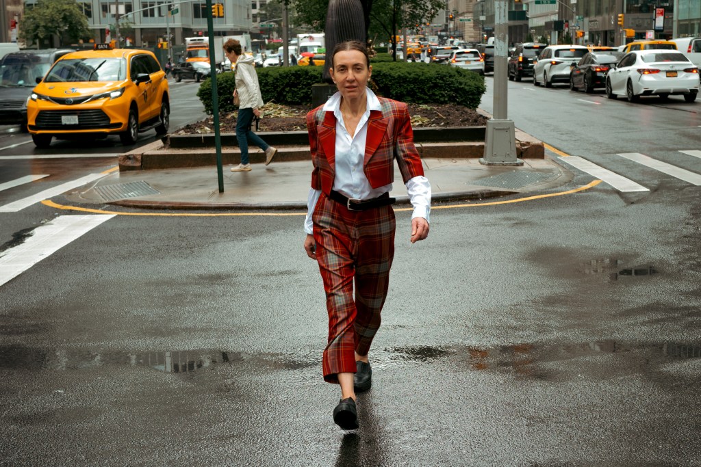 A person wearing a stylish plaid suit confidently walks through a busy urban street with cars and pedestrians in the background.