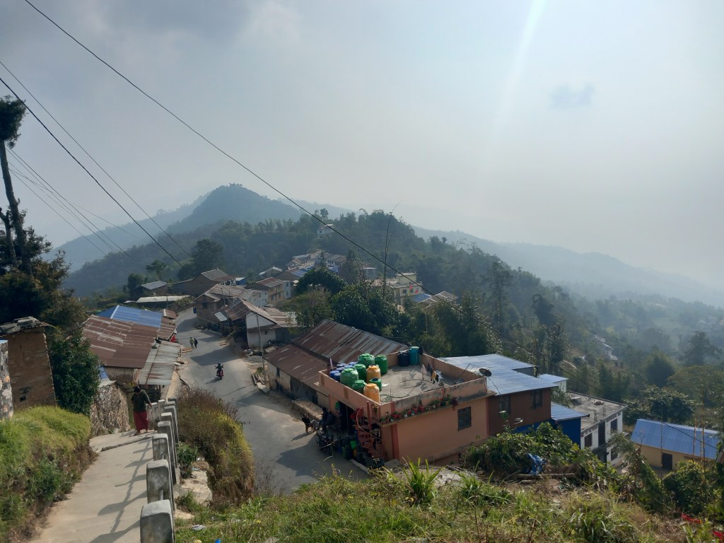 A view of the village of Pokhare in Nepal, showcasing small houses with tin roofs nestled among hills and misty mountains in the background.