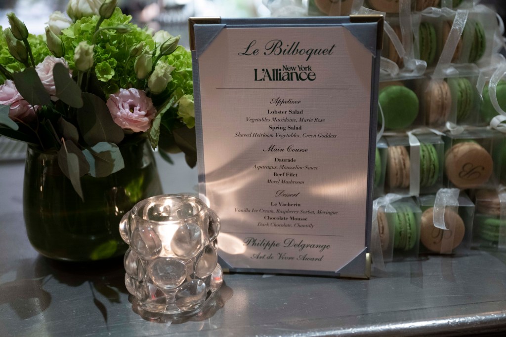 A beautifully arranged table featuring a floral centerpiece, a lit candle, and a menu for Le Bilboquet at L'Alliance New York, highlighting the event menu including appetizers, main courses, and desserts.