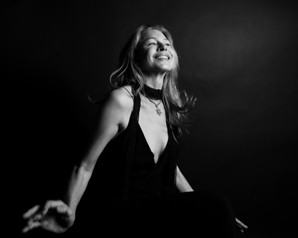 A black-and-white portrait of a woman with long hair, smiling joyfully while seated against a dark background.