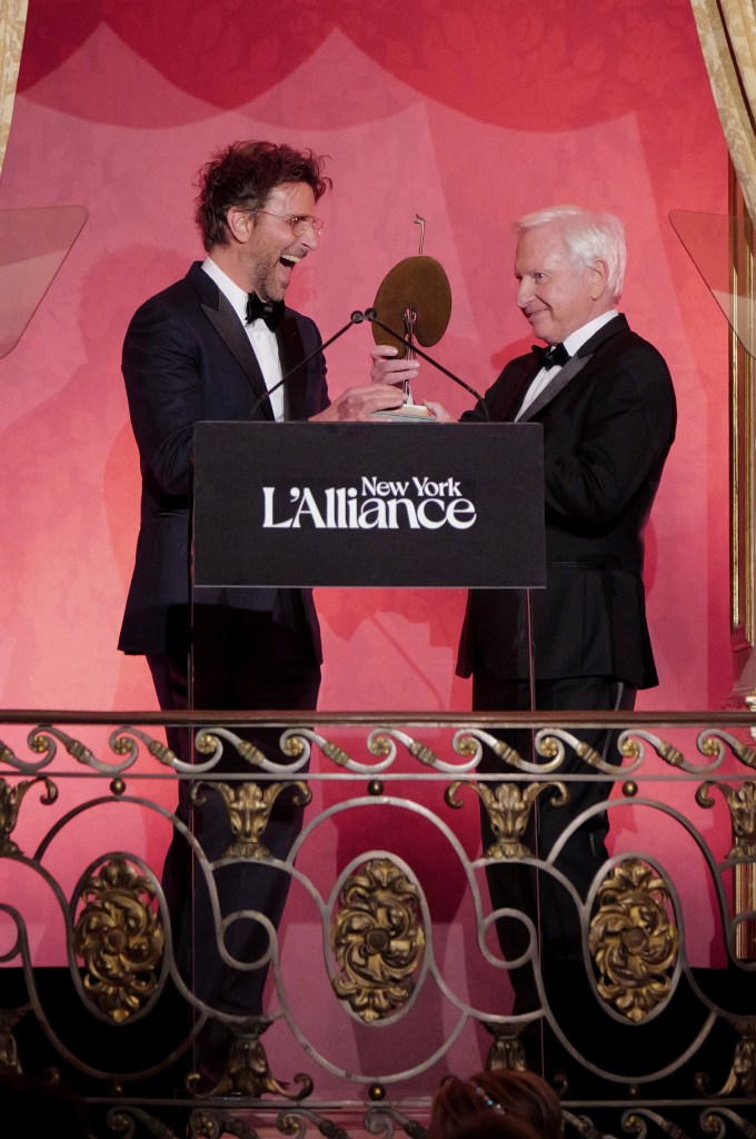 Two men in formal attire share a joyful moment at the L'Alliance New York Gala, one presenting an award while the other accepts it, with an elegant backdrop.
