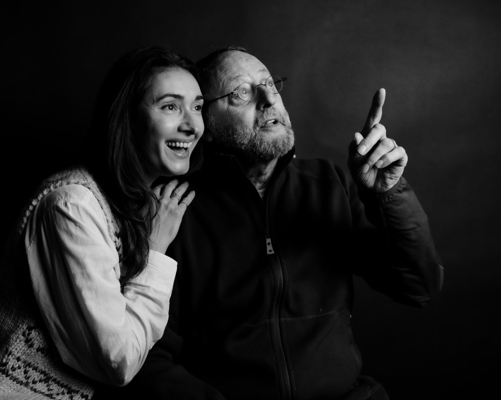 A black-and-white portrait of a man and a woman sharing a joyful moment, with the woman laughing and resting her hand on the man's shoulder as he points upward, suggesting a sense of wonder and connection.