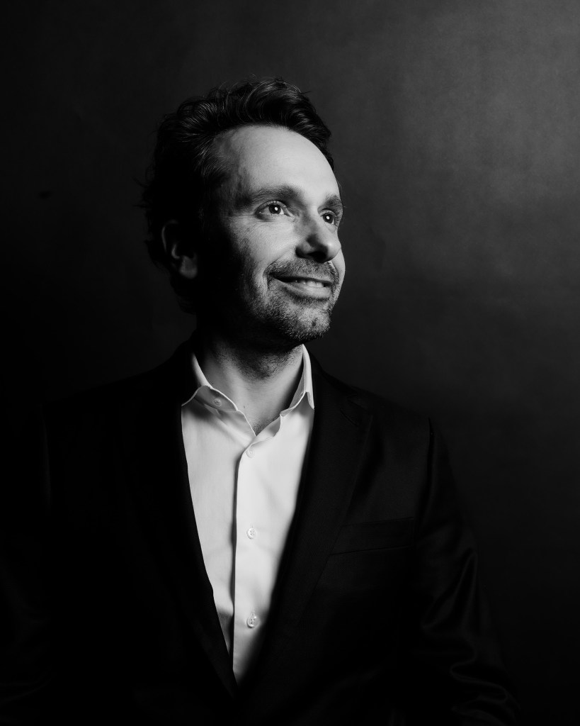 Black-and-white portrait of a man in a suit, smiling slightly with a thoughtful expression against a dark background.