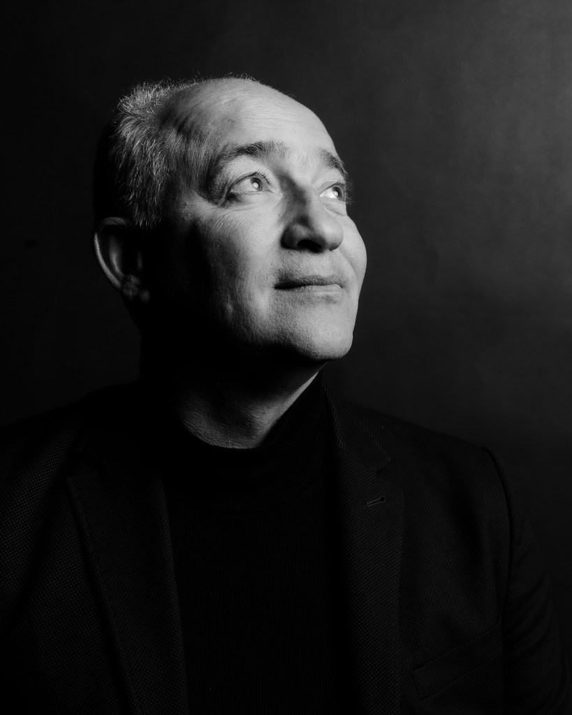 A black-and-white portrait of a man looking upward with a thoughtful expression, dressed in a dark blazer and turtleneck against a plain dark background.