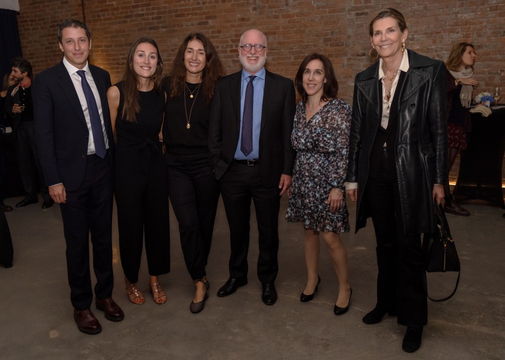 A group of six individuals poses for a photo at a fundraising event, in front of a brick wall. They are dressed in formal attire, smiling and engaging with the camera.