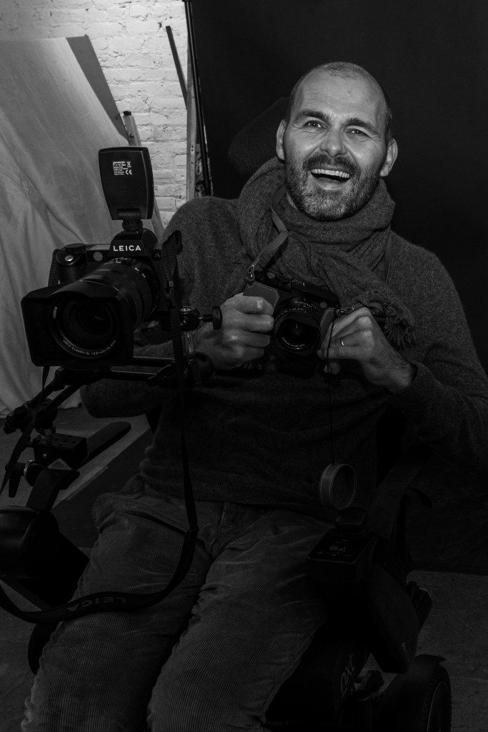 A black and white portrait of a smiling man in a wheelchair, holding two cameras, showcasing a joyful expression.