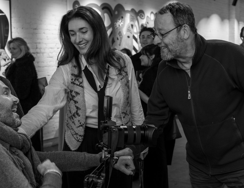 A black and white photo of three people interacting warmly in an art gallery setting, with one person in a wheelchair smiling and engaging with a woman and another man, showcasing a sense of community and support.