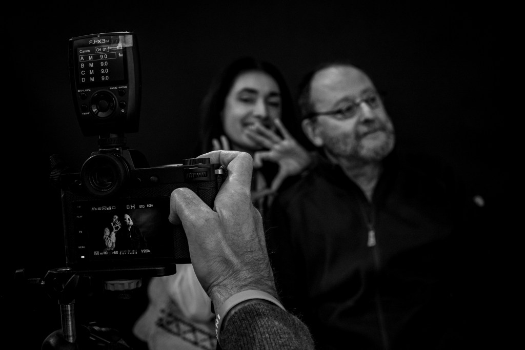 A close-up of a camera displaying a photo of two people posing, with one person smiling and making a hand gesture. The image captures the photographer's hand adjusting the camera settings, with a dark background.