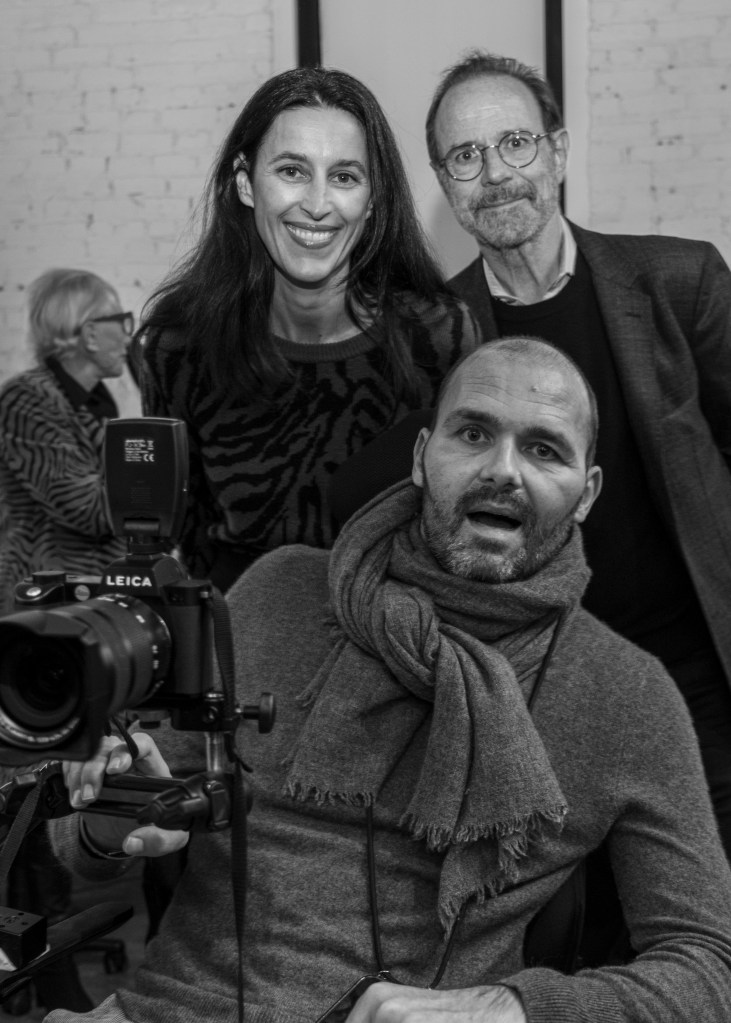 A black-and-white portrait of three individuals in a casual setting. A woman smiles in the foreground, while a man sits in a wheelchair with a camera. Another man stands behind them, looking towards the camera.