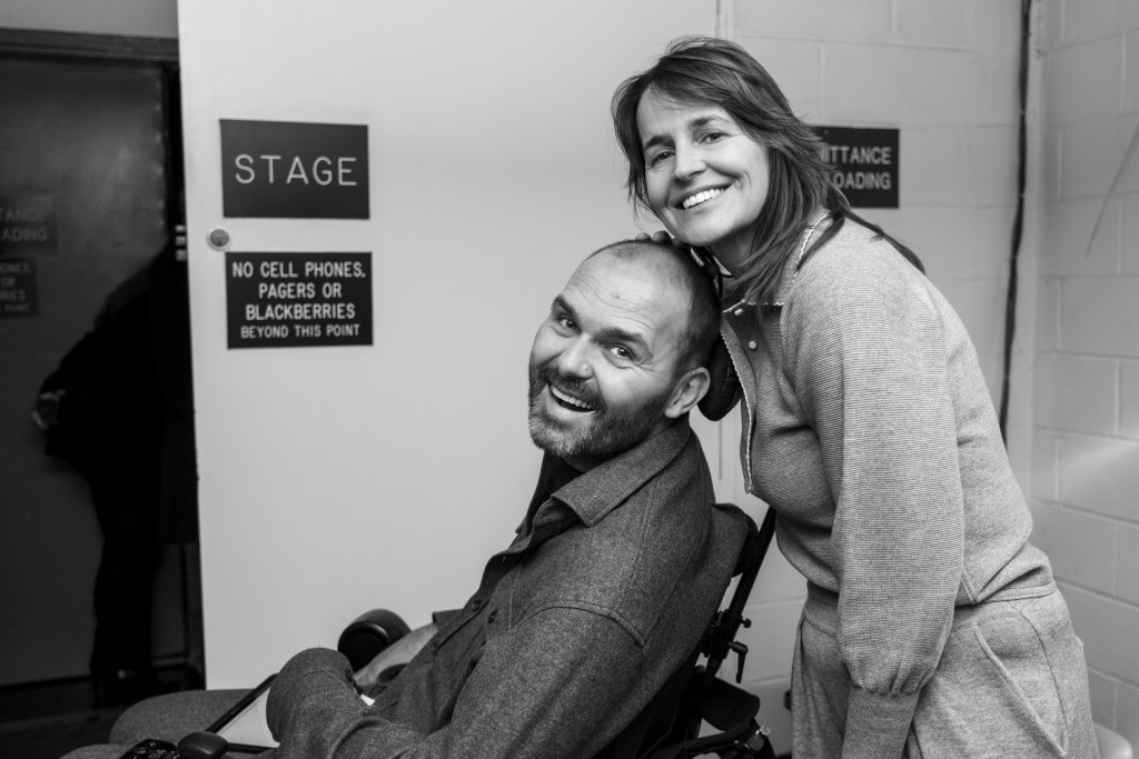 A smiling man in a wheelchair poses with a woman in casual attire, both appearing joyful against a backdrop of a door marked 'STAGE'.