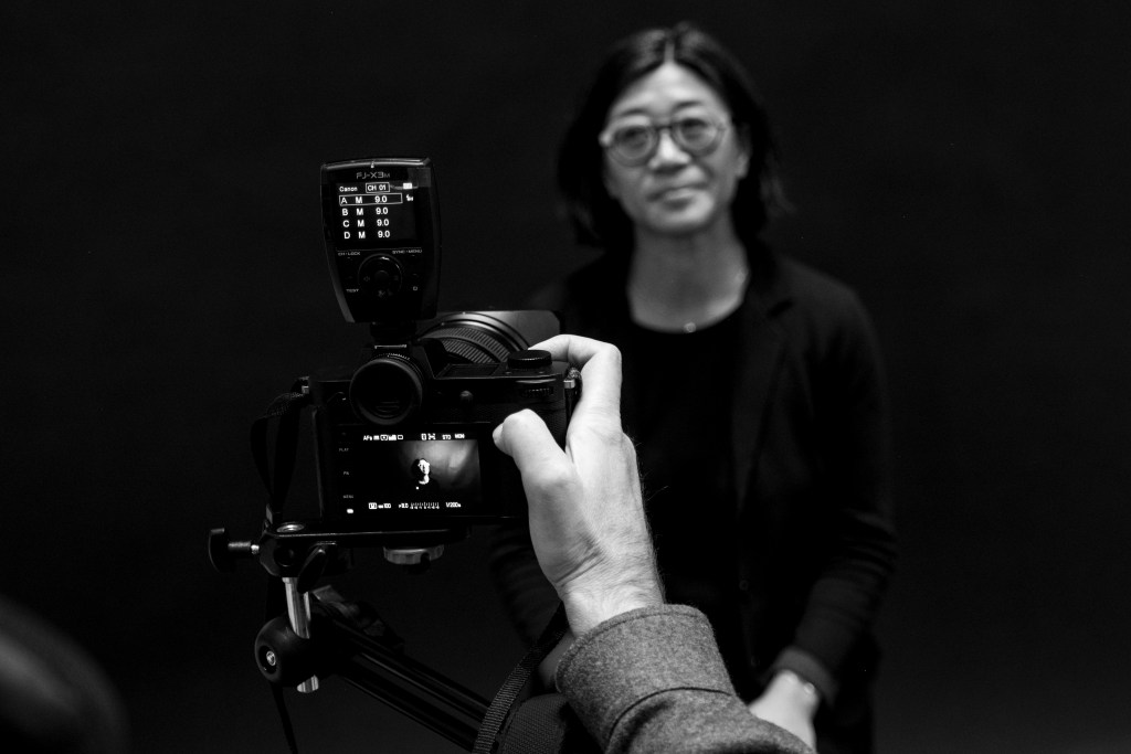 A photographer captures a portrait of a woman in front of a black backdrop during a photoshoot.