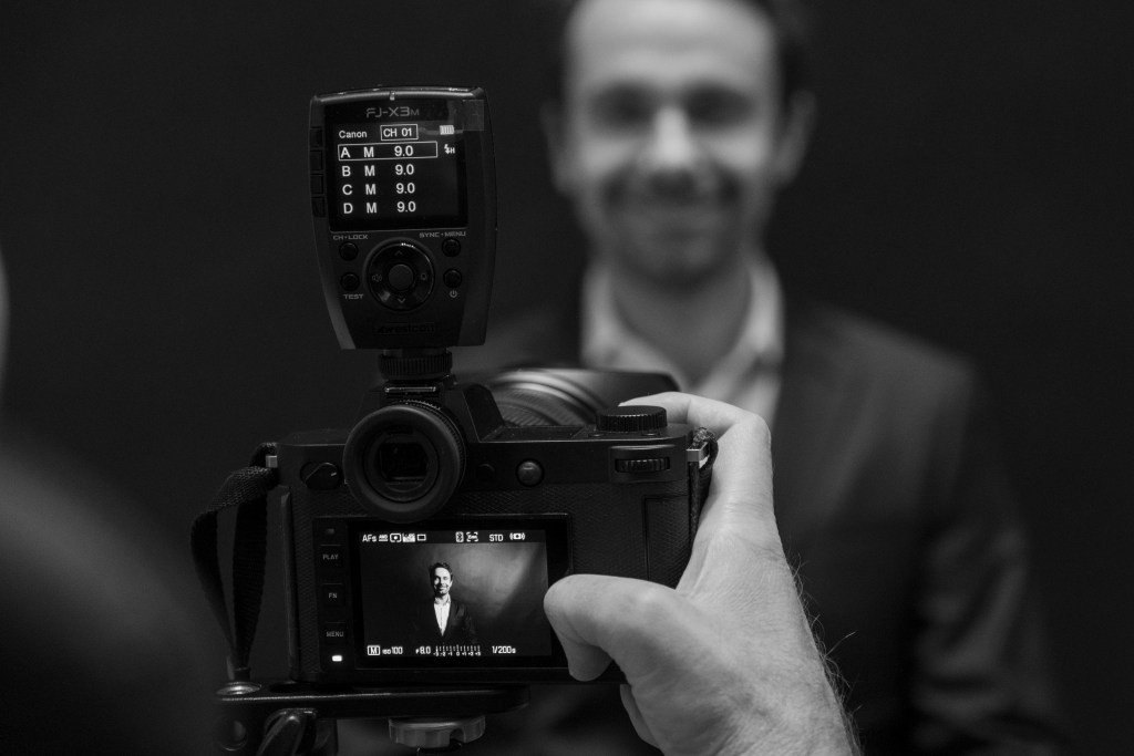 A black-and-white photograph showing a camera focused on a smiling man in a suit, with a photographer's hand visible holding the camera, suggesting a portrait session.