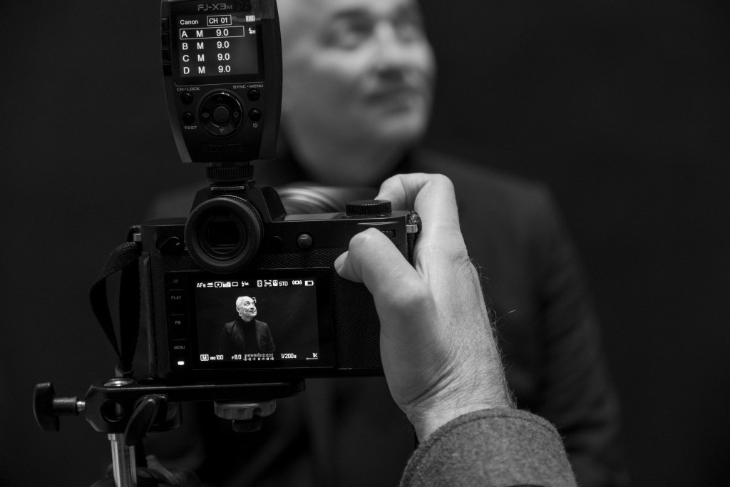 A photographer taking a black-and-white portrait of a smiling subject, with camera settings visible on the flash unit.