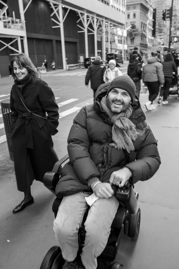 Black-and-white image of a man in a wheelchair, smiling and wearing a winter coat and a scarf, on a busy city street, while a woman walks beside him, both appearing joyful.