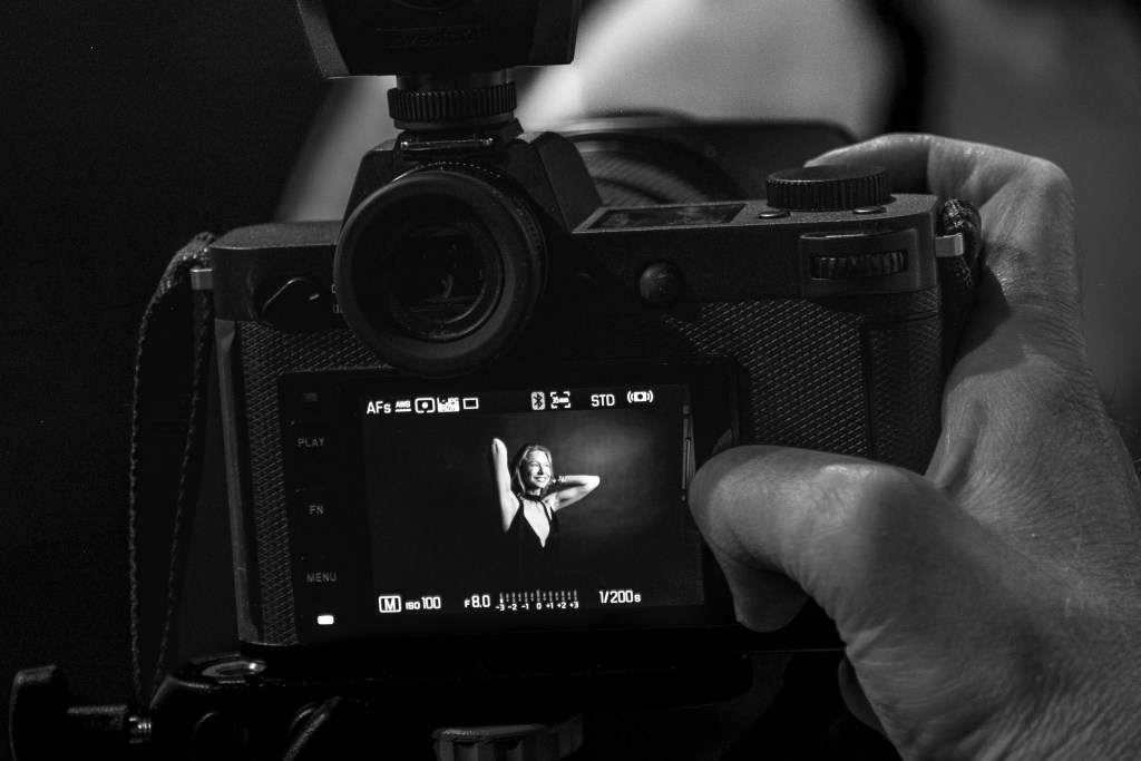 A close-up shot of a hand holding a camera, capturing a woman posing in front of the lens, visible on the camera screen. The image is in black-and-white, emphasizing the artistic and candid nature of the moment.