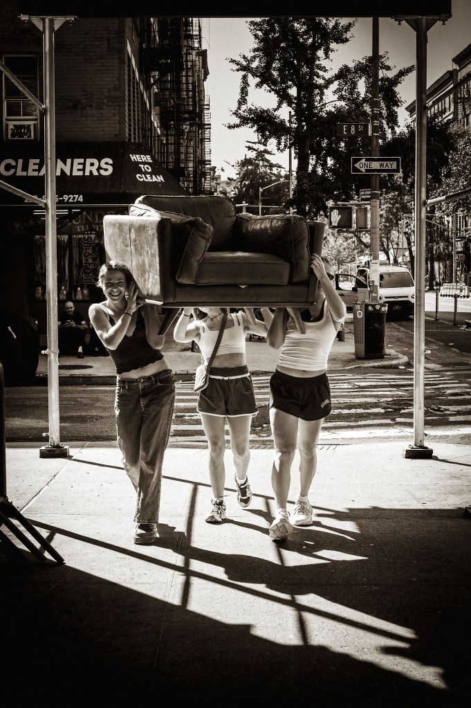 Three people carrying a couch along a street in New York City, with trees and buildings in the background, in black and white.