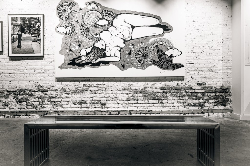 A black and white photograph of an art gallery featuring a large mural of a reclining figure surrounded by whimsical elements, with a metal bench in the foreground and smaller framed photographs displayed on the wall.