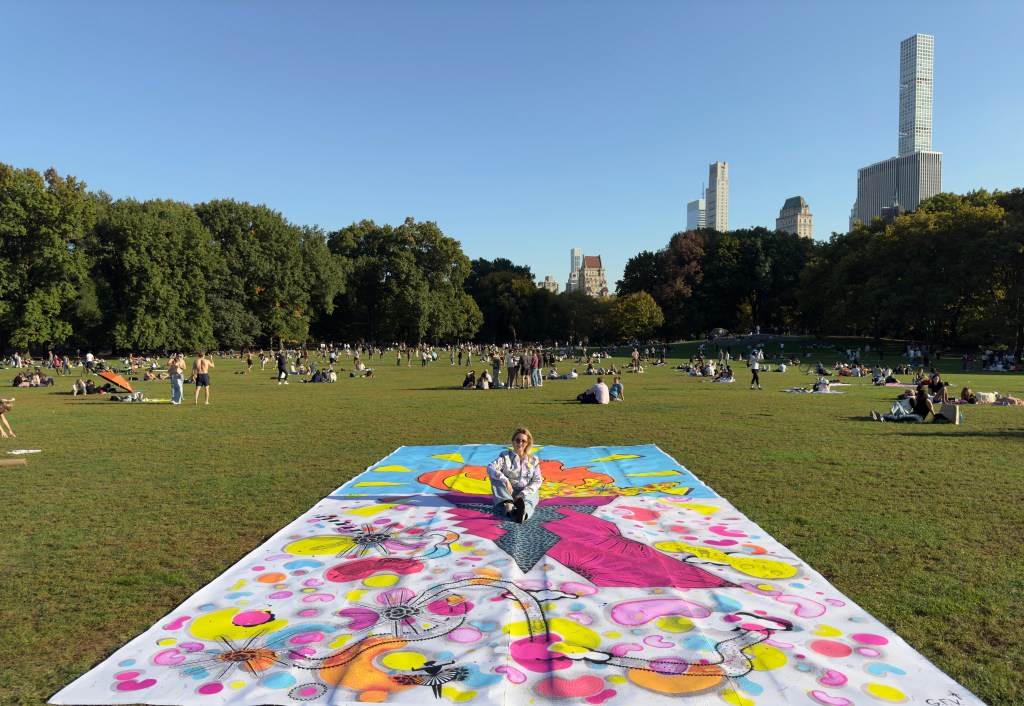 An artist sitting on a colorful mural in Central Park, surrounded by people on the grass and tall buildings in the background, showcasing a vibrant urban art piece.