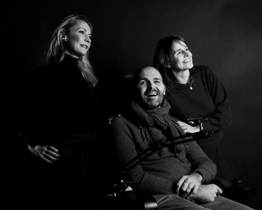 A black and white portrait of three individuals, two women standing beside a man in a wheelchair, all smiling while looking in different directions against a dark background.