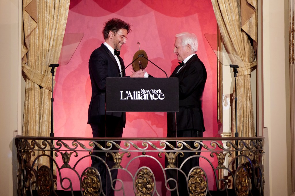 Two men in formal attire on a balcony stage, one presenting an award to the other, with a backdrop displaying 'New York L'Alliance.'