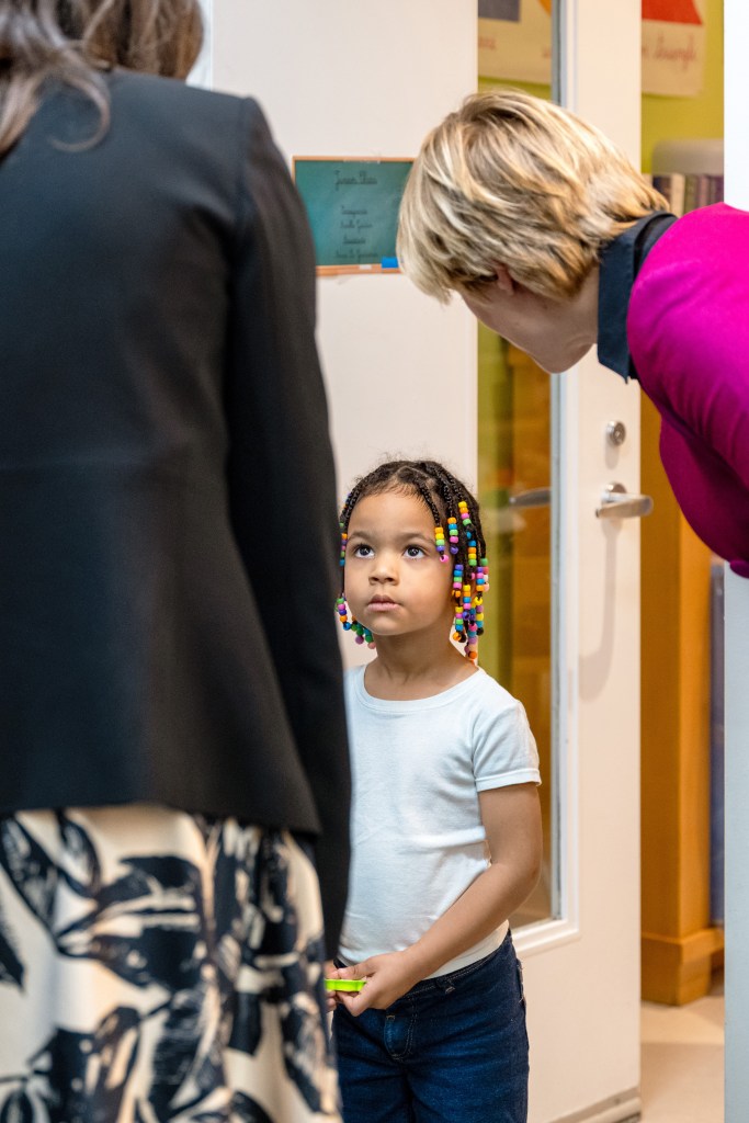 A young girl with colorful beads in her hair looking up at two adults in conversation.
