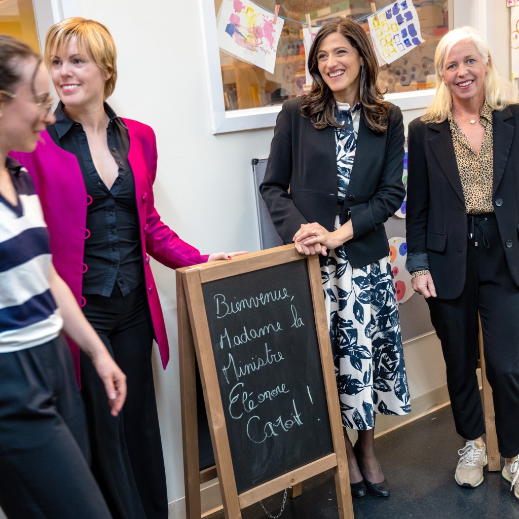 A group of four women standing together, smiling, near a chalkboard sign that says 'Bienvenue, Madame la Ministre Éducation Carole!' with colorful artwork displayed in the background.