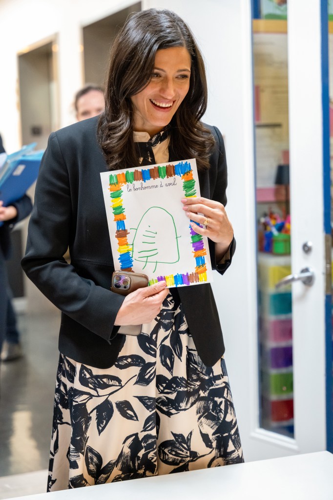 A woman holding a colorful drawing created by a child, smiling happily while wearing a black blazer and floral dress.