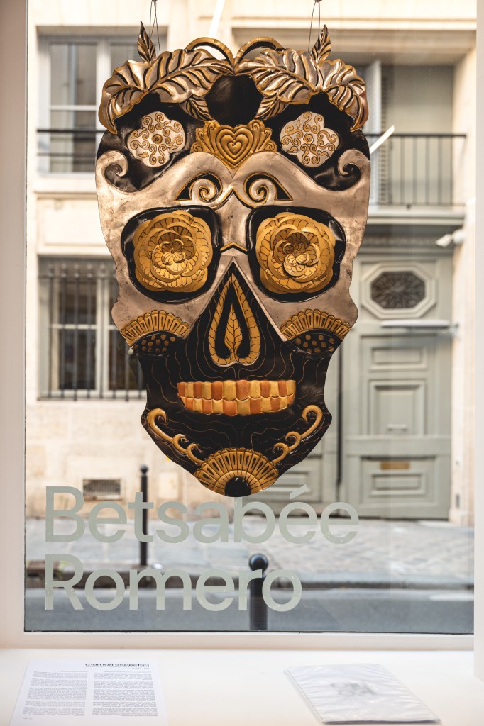 A decorative skull art piece by Betsabé Romero, featuring intricate designs and vibrant colors, displayed in a window with street view behind it.
