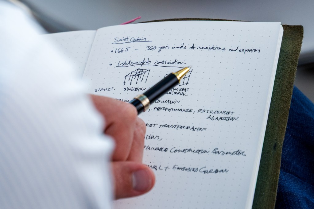 A close-up of a person's hand holding a pen, writing in a notebook filled with handwritten notes and sketches about construction and structural design.