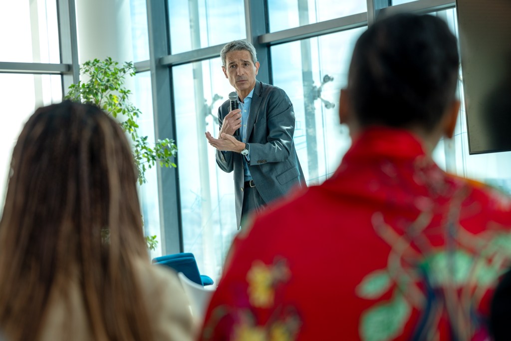 A businessman speaking to a small audience in a modern office setting, with large windows and plants in the background.