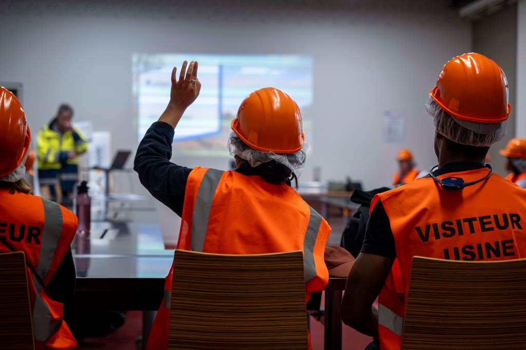 A group of people in safety gear, including orange vests and hard hats, are seated in a classroom setting, with one person raising their hand to ask a question. A presentation is visible on the screen in the background.