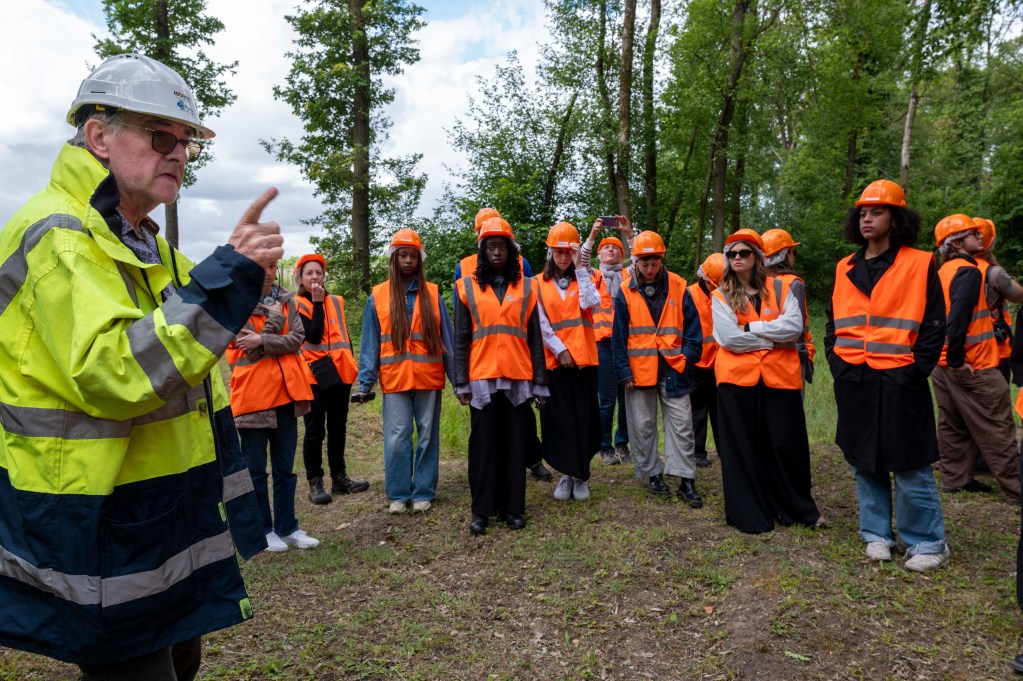 An older man in a yellow and blue reflective jacket and a hard hat gestures while speaking to a group of people wearing orange safety vests and helmets in a wooded area.