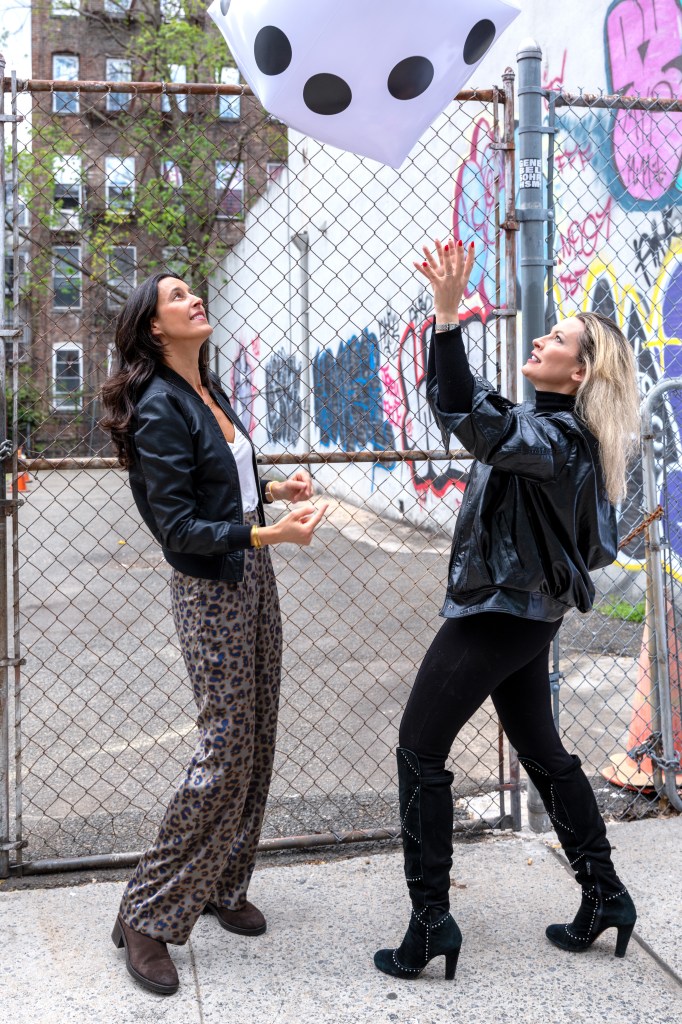 Two women playfully interacting with a large inflatable dice in an urban setting, surrounded by graffiti and a chain-link fence.