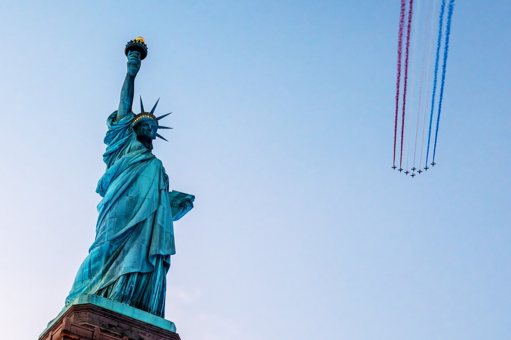 The Statue of Liberty towering against the sky with an aircraft formation trailing red, white, and blue smoke above.