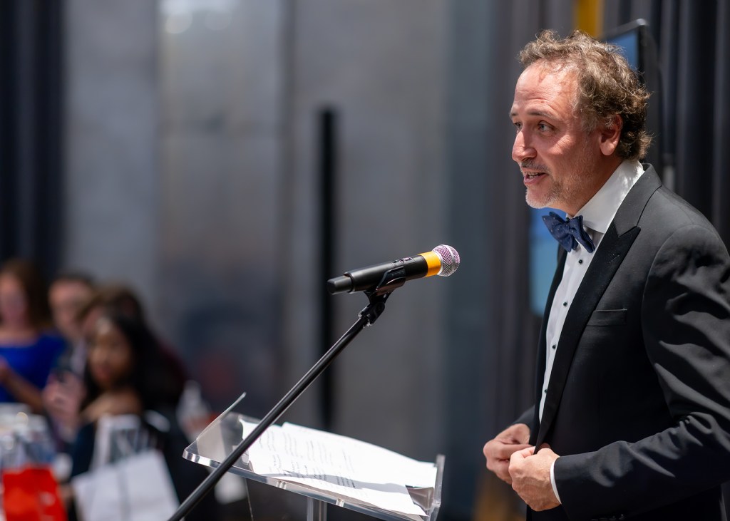 A man in a tuxedo with a bow tie speaks at a podium with a microphone in a formal setting, while an audience is seated in the background.