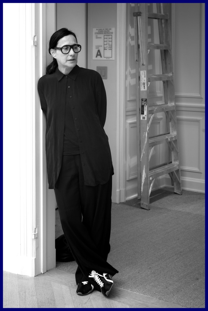 A woman in black attire stands casually in an interior space, leaning against a doorframe with a ladder visible in the background.