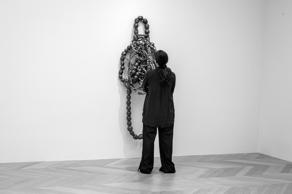 A person standing in front of a large, abstract sculpture made of intertwined black objects, with a minimalist white wall in the background.